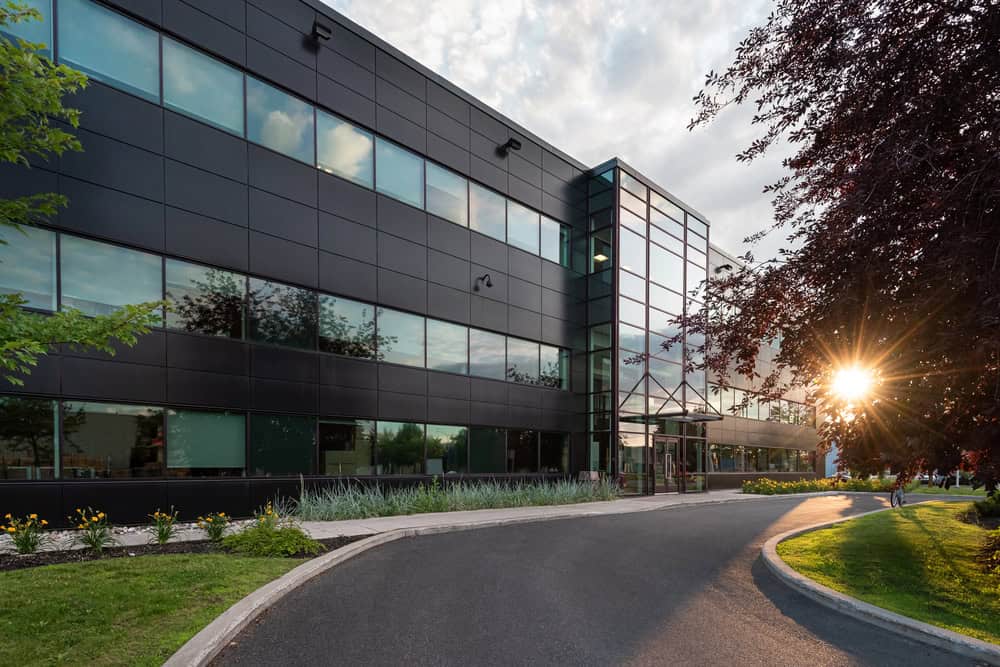 Modern office building with large windows and dark exterior panels, surrounded by professional grounds maintenance and commercial landscaping in Tarrant County, at sunset with sunlight filtering through trees.