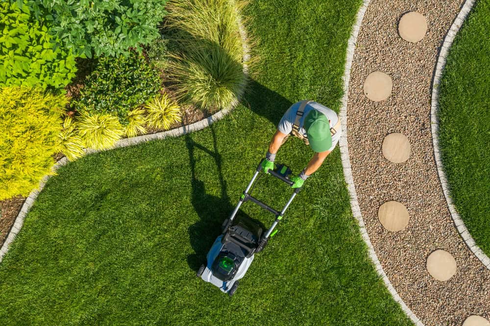 Person mowing a well-manicured lawn with a push mower, surrounded by landscaped plants and a decorative stone pathway, showcasing expert lawn care Tarrant County.